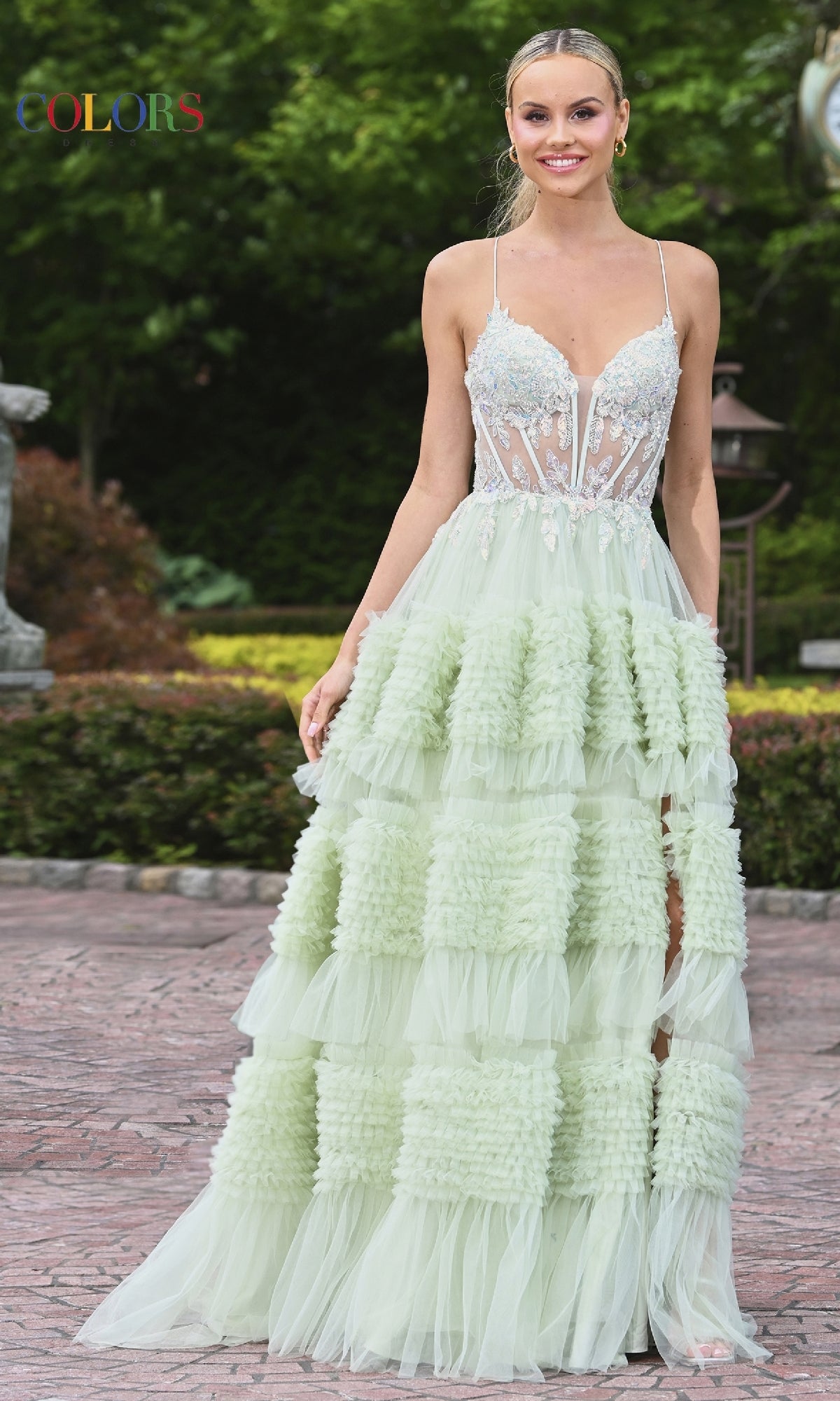 A woman stands outdoors in the Mini-Ruffle Long Prom Ball Gown: Colors Dress 3515, featuring a pale green hue, ruffled skirt, sheer lace-embellished bodice, and spaghetti straps. Green trees and stone pavement are in the background.